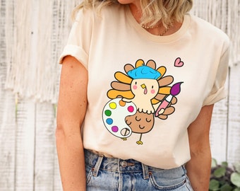 May include: A beige t-shirt with a cartoon turkey wearing a blue beret and holding a paintbrush and a paint palette. The turkey is surrounded by colorful paint splatters. The text "Natural" is visible at the bottom of the image.