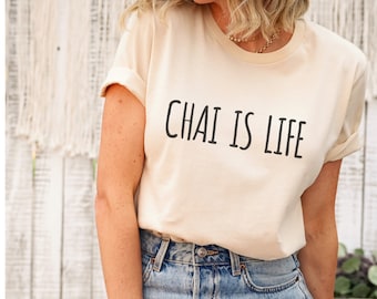 Może przedstawiać: Kobieta w jasnobeżowej koszulce z nadrukiem "Chai is Life" w kolorze czarnym. Ma na sobie niebieskie jeansy z przetarciami.