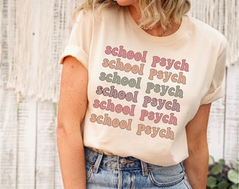 May include: A light beige t-shirt with the text "school psych" repeated six times in a retro font. The text is in shades of pink, peach, and green.