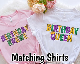 May include: Two matching t-shirts, one pink with the text "Birthday Krewe" in green, purple, and yellow, and one white with the text "Birthday Queen" in green, purple, and yellow with a gold crown above the word "Queen".