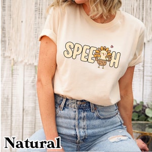 Peut inclure: Un t-shirt beige avec le mot "SPEECH" imprimé dessus dans une police de caractères de dessin animé. Une dinde de dessin animé se tient au milieu du mot. La dinde est brune et orange avec un cœur rouge au-dessus.
