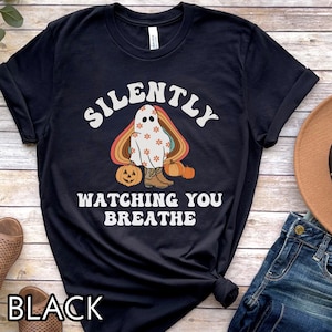 May include: Black t-shirt with a retro-style graphic of a ghost wearing cowboy boots and holding pumpkins. The text "Silently Watching You Breathe" is printed on the shirt.