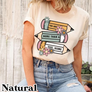 May include: A beige t-shirt with a graphic of three pencils with flowers. The pencils have the text "You are more than a test score" on them.