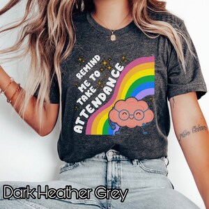 May include: Dark heather grey t-shirt with a rainbow and a cartoon brain wearing glasses. The text on the shirt reads "Remind me to take attendance".