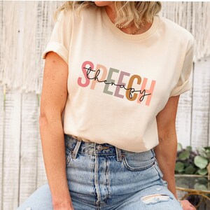 May include: A light beige t-shirt with the text "SPEECH therapy" printed in rainbow colors. The text is in a stylized font with each letter in a different color.