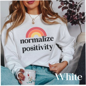 May include: White sweatshirt with a rainbow graphic and the text "normalize positivity" printed on the front.