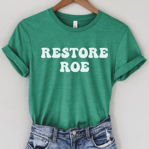 May include: Green t-shirt with white text that reads "Restore Roe" in a retro font.