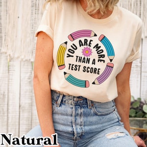 May include: A natural colored t-shirt with a graphic design of colorful pencils in a circle around the text "You are more than a test score".