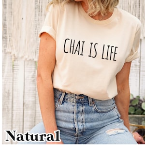 May include: A woman wearing a light beige t-shirt with the text "Chai is Life" printed in black. She is wearing blue jeans with a distressed look.