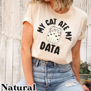 May include: A beige t-shirt with a black graphic of a cat wearing glasses and the text "My Cat Ate My Data".