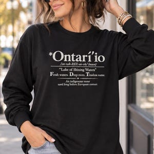 May include: A black long-sleeve sweatshirt featuring white text that reads *Ontari'io, on-tah-REE-ee-oh (noun), Lake of Shining Waters, Fresh waters. Deep roots. Timeless name. An indigenous name used long before European contact.