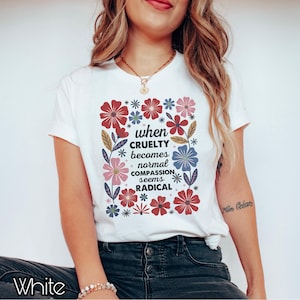 May include: White t-shirt with a floral design and the text "when CRUELTY becomes normal COMPASSION seems RADICAL". The flowers are red, pink, and blue. The shirt is a casual wear item.
