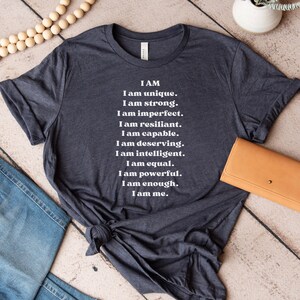 May include: A dark gray t-shirt with white text that reads "I AM I am unique. I am strong. I am imperfect. I am resilient. I am capable. I am deserving. I am intelligent. I am equal. I am powerful. I am enough. I am me."