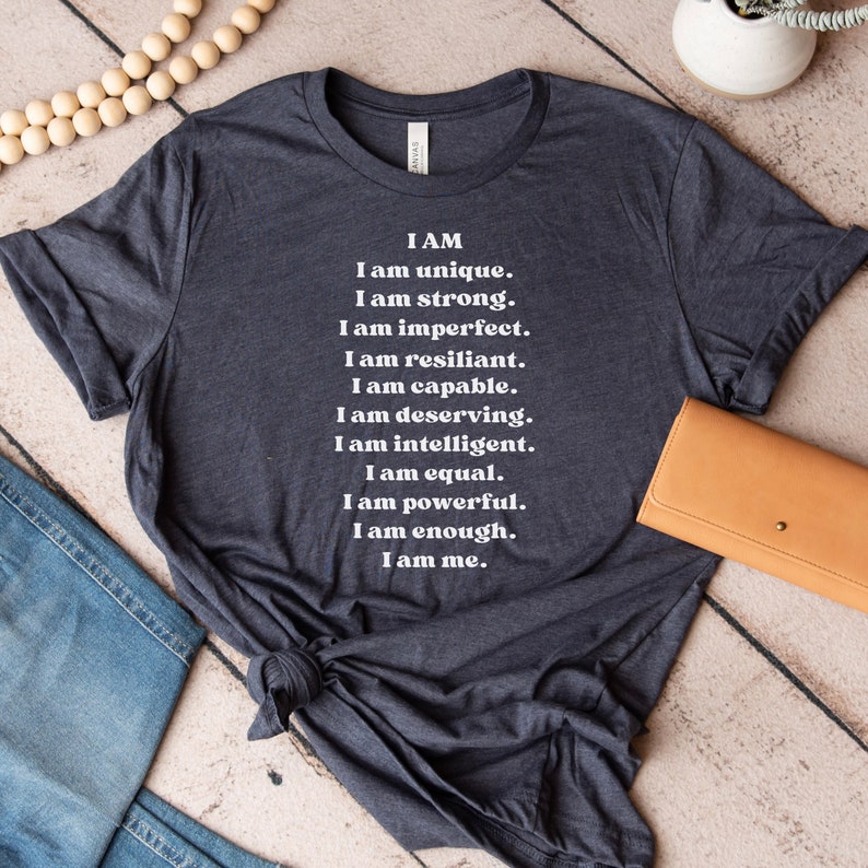May include: A dark gray t-shirt with white text that reads "I AM I am unique. I am strong. I am imperfect. I am resilient. I am capable. I am deserving. I am intelligent. I am equal. I am powerful. I am enough. I am me."