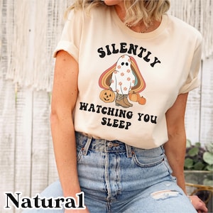 Peut inclure: Un t-shirt crème avec un graphique rétro d'un fantôme portant des bottes de cowboy et tenant une citrouille. Le texte "Silently Watching You Sleep" est imprimé sur le t-shirt.
