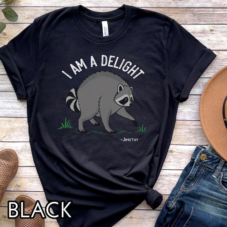 May include: A black short-sleeve t-shirt featuring a graphic of a gray raccoon with a striped tail. The text above the raccoon reads I AM A DELIGHT in white capital letters, and the name -JIMOTHY is printed in white below the raccoon.