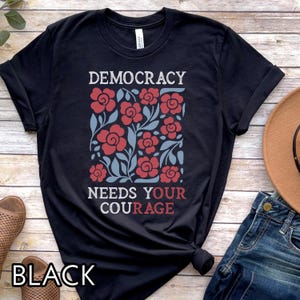 May include: Black t-shirt with the words "DEMOCRACY NEEDS YOUR COURAGE" in white and red. The shirt features a floral design with red flowers and blue leaves. The word "BLACK" is at the bottom.