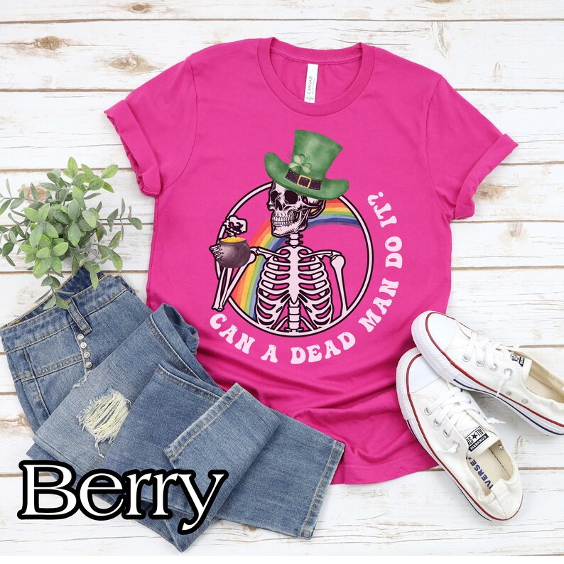 May include: A pink t-shirt with a graphic of a skeleton wearing a green leprechaun hat and holding a pot of gold. The text on the shirt reads "Can a dead man do it?"