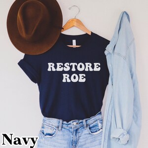 May include: A navy blue t-shirt with the text "RESTORE ROE" in white, retro-style lettering. The shirt is hanging on a wooden hanger and is styled with a brown hat and a light blue denim jacket.