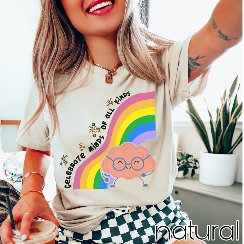 Pu&ograve; includere: Una maglietta bianca con un arcobaleno e un cervello di cartone animato con gli occhiali. Il testo sulla maglietta dice "Celebrate Minds of All Kinds".
