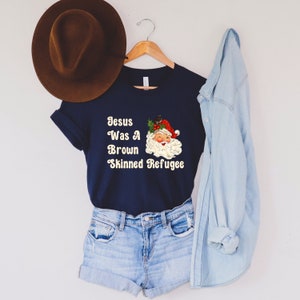 May include: A navy blue t-shirt with a white graphic of a cartoon Santa Claus with the text "Jesus Was A Brown Skinned Refugee".