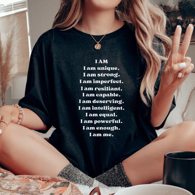 May include: A black t-shirt with white text that reads "I AM I am unique. I am strong. I am imperfect. I am resilient. I am capable. I am deserving. I am intelligent. I am equal. I am powerful. I am enough. I am me."