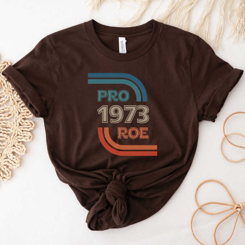 May include: Brown t-shirt with a retro-style graphic that says "PRO 1973 ROE" in blue, orange, and white.