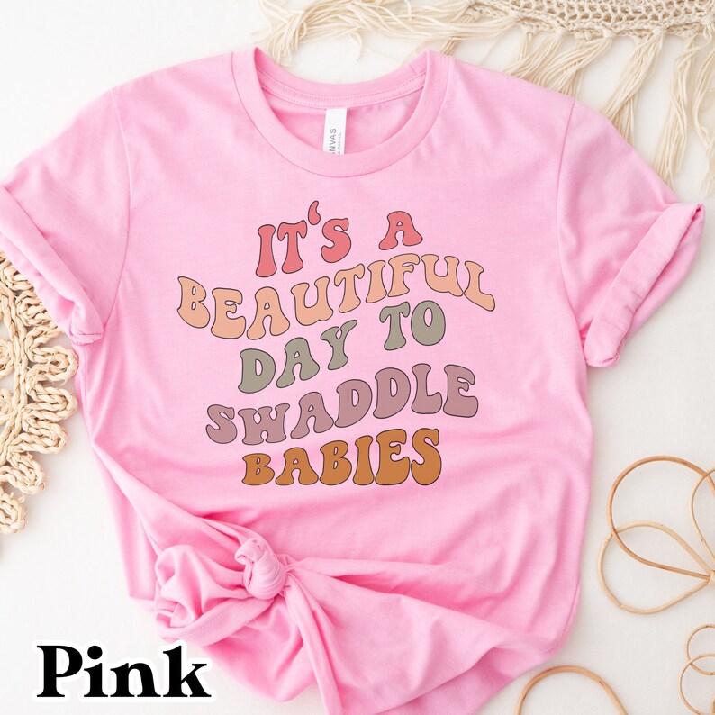 K&ouml;nnte beinhalten: Rosa T-Shirt mit dem Text "It's a beautiful day to swaddle babies" in einer Retro-Schriftart mit bunten Buchstaben.