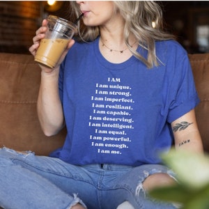 May include: A blue t-shirt with white text that reads "I AM I am unique. I am strong. I am imperfect. I am resilient. I am capable. I am deserving. I am intelligent. I am equal. I am powerful. I am enough. I am me."