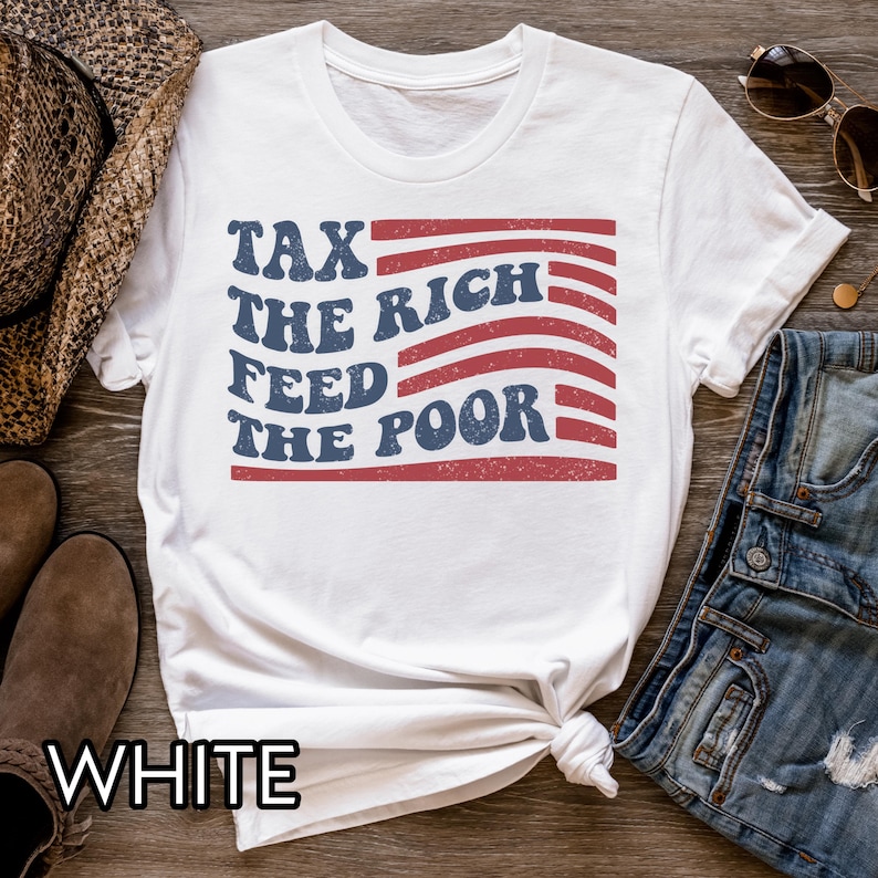 May include: White t-shirt with the text "TAX THE RICH FEED THE POOR" in blue and red, designed to resemble a flag. The shirt is styled with rolled-up sleeves and a knot at the waist. Sunglasses and denim jeans are visible.