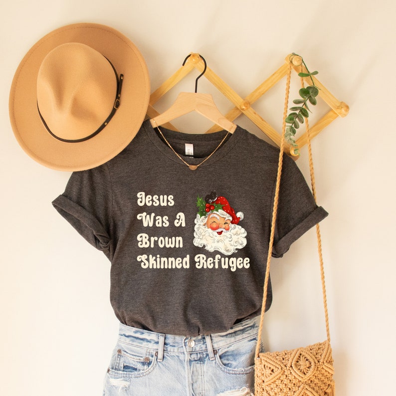 May include: A dark grey t-shirt with a white graphic that says "Jesus Was A Brown Skinned Refugee" and a cartoon image of Santa Claus.
