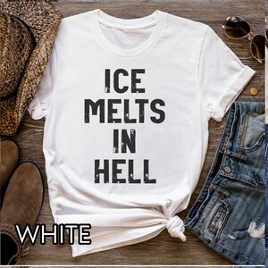 May include: White t-shirt with the text "ICE MELTS IN HELL" in black, distressed font. The word "WHITE" is at the bottom of the shirt. The shirt is knotted at the bottom. Sunglasses and a cowboy hat are in the background.