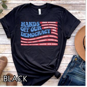 May include: Black t-shirt with a red, white, and blue American flag design. The text "Hands Off Our Democracy" is printed in blue with the words "Free Speech", "Federal Programs", "Healthcare", "DEI", "Students", "Medicaid", "Medicare", "Federal Workers", "Scientists", "National Parks", "Courts", "Immigrants", "Personal Data", "Our Rights", "Fair Elections", "Education", and "Social Security" printed in red.