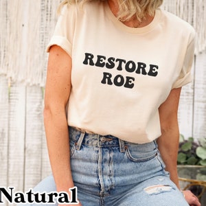 May include: A woman wearing a natural colored t-shirt with the text "RESTORE ROE" in black retro-style lettering. She is wearing blue jeans.