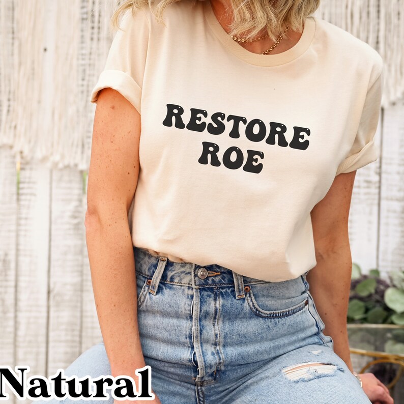 May include: A woman wearing a natural colored t-shirt with the text "RESTORE ROE" in black retro-style lettering. She is wearing blue jeans.