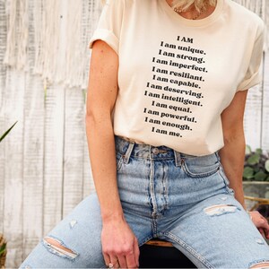 May include: A white t-shirt with the text "I AM" and a list of positive affirmations, including "unique", "strong", "resilient", "capable", "deserving", "intelligent", "equal", "powerful", "enough", and "me".