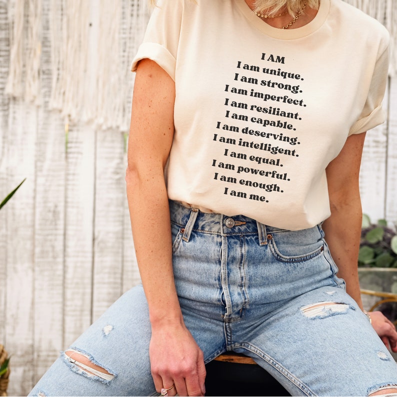 May include: A white t-shirt with the text "I AM" and a list of positive affirmations, including "unique", "strong", "resilient", "capable", "deserving", "intelligent", "equal", "powerful", "enough", and "me".