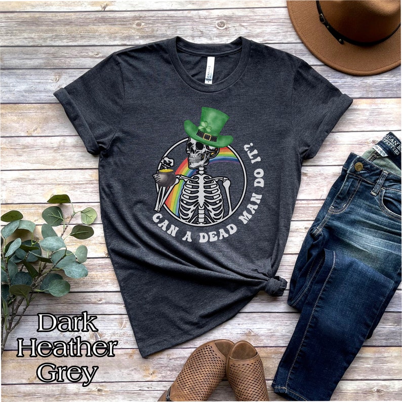 May include: Dark heather grey t-shirt with a skeleton wearing a green leprechaun hat and holding a pot of gold. The text on the shirt reads "Can a dead man do it?"
