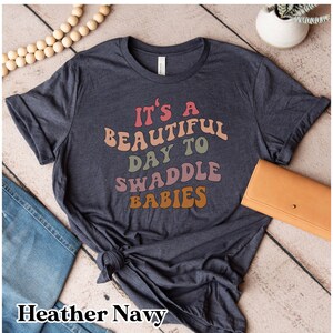 K&ouml;nnte beinhalten: Ein heidefarbenes marineblaues T-Shirt mit dem Text "Es ist ein wundersch&ouml;ner Tag zum Wickeln von Babys" in Regenbogenfarben.