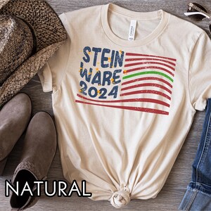 May include: A beige t-shirt with a distressed American flag design and the text "STEIN WARE 2024" printed on the front. The shirt is made of a soft, comfortable material and has a natural color.
