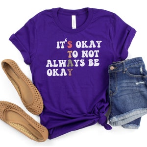 May include: Purple t-shirt with the text "It's okay to not always be okay" in rainbow colored letters.