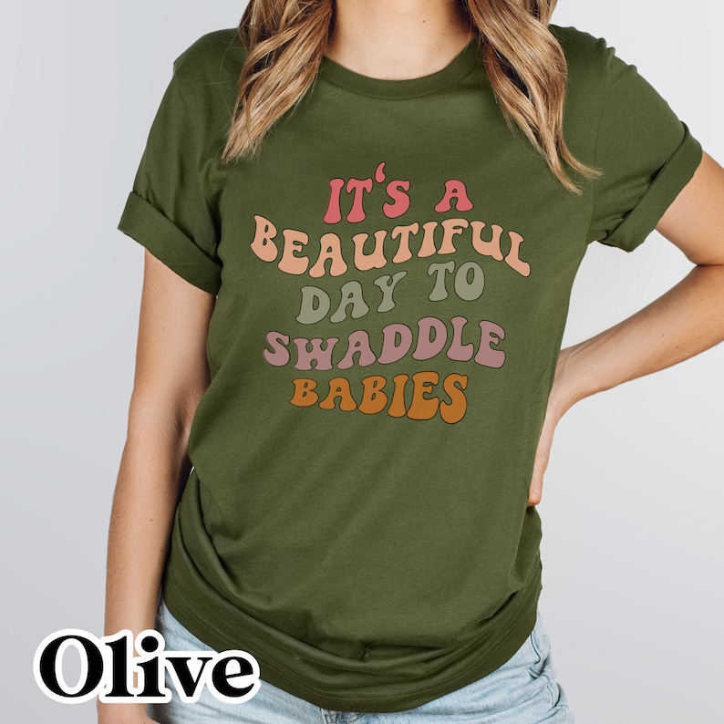 K&ouml;nnte beinhalten: Olivengr&uuml;nes T-Shirt mit einer Retro-Grafik, auf der "Es ist ein wundersch&ouml;ner Tag, um Babys zu wickeln" steht.