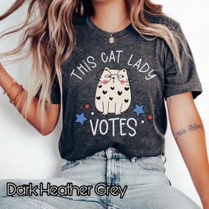 May include: Dark heather grey t-shirt with a cartoon cat wearing glasses and the text "This Cat Lady Votes" with red and blue stars.