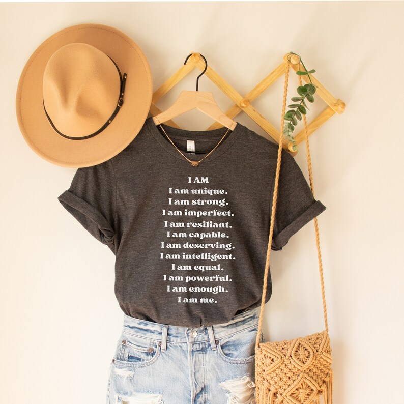 May include: A dark grey t-shirt with white text that reads "I AM I am unique. I am strong. I am imperfect. I am resilient. I am capable. I am deserving. I am intelligent. I am equal. I am powerful. I am enough. I am me."