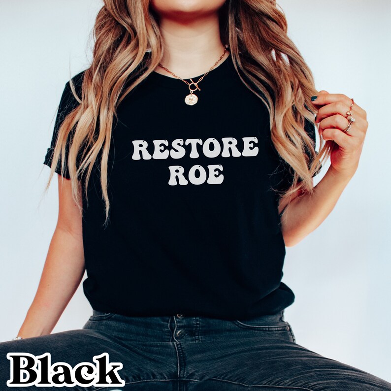 May include: Black t-shirt with white text that reads "Restore Roe" in a retro font.