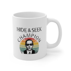May include: White ceramic mug with a vintage-style graphic of a man with glasses and the text "HIDE & SEEK CHAMPION" in black.
