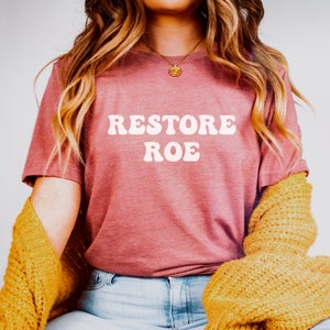May include: A woman wearing a mauve t-shirt with the text "RESTORE ROE" in white retro-style lettering. She is wearing blue jeans and a yellow cardigan.