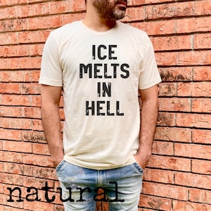May include: Cream-colored t-shirt with the text "ICE MELTS IN HELL" in bold, black letters. The shirt is worn by a person in blue jeans, standing in front of a brick wall. The word "natural" is printed at the bottom of the image.