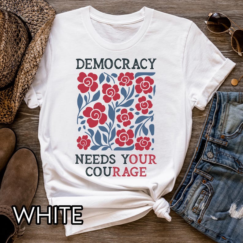 May include: White t-shirt with the words "DEMOCRACY NEEDS YOUR COURAGE" in black and red. The shirt features a floral design with red roses and blue leaves. The word "WHITE" is printed at the bottom.