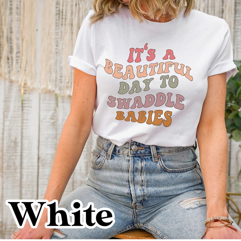 K&ouml;nnte beinhalten: Wei&szlig;es T-Shirt mit einer Retro-Grafik, auf der "Es ist ein sch&ouml;ner Tag, um Babys zu wickeln" steht.